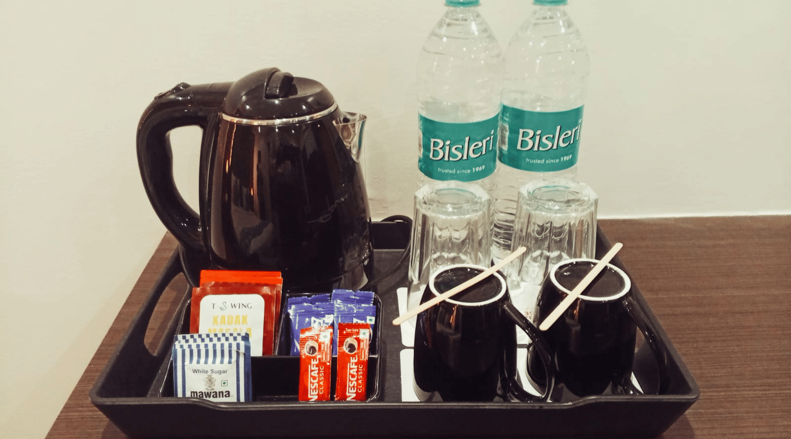 A hospitality station at Hotel Mumbai House, Thane featuring a black electric kettle, 2 plastic water bottles, 2 ceramic mugs, and various tea sachets arranged neatly on a rectangular tray.