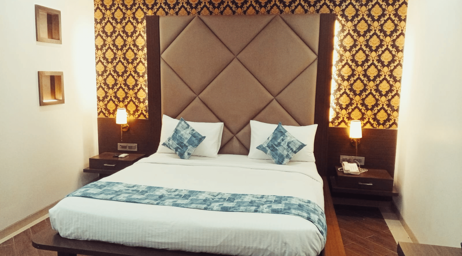 A symmetrical view of a king size bed set against a tall patterned wallpaper headboard with 2 bedside drawers and small wall frames at Hotel Mumbai House, Thane.