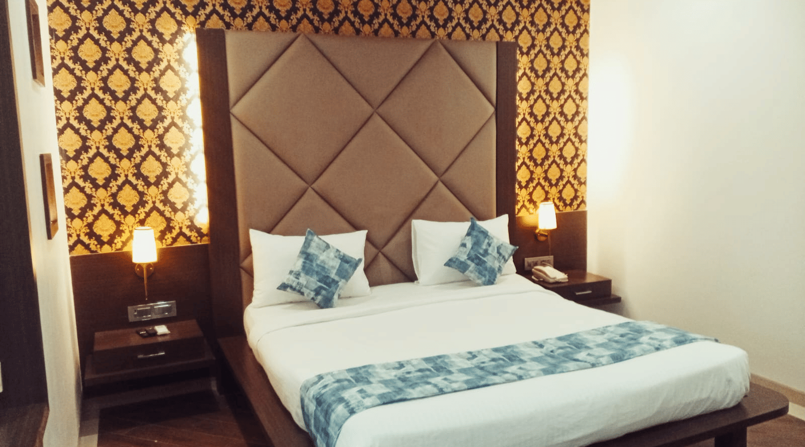 A king size bed with a padded headboard, a patterned wallpaper background, 2 bedside tables, and 2 illuminated wall lamps at Hotel Mumbai House, Thane.