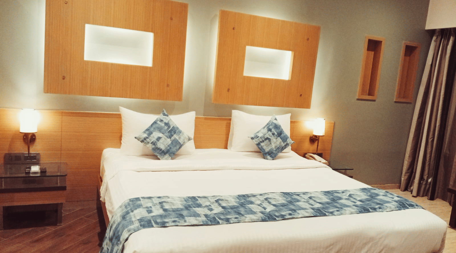 A king size bed with a white duvet, a patterned bed runner, 4 pillows, 2 decorative cushions, and 2 rectangular wall headboard panels at Hotel Mumbai House, Thane.