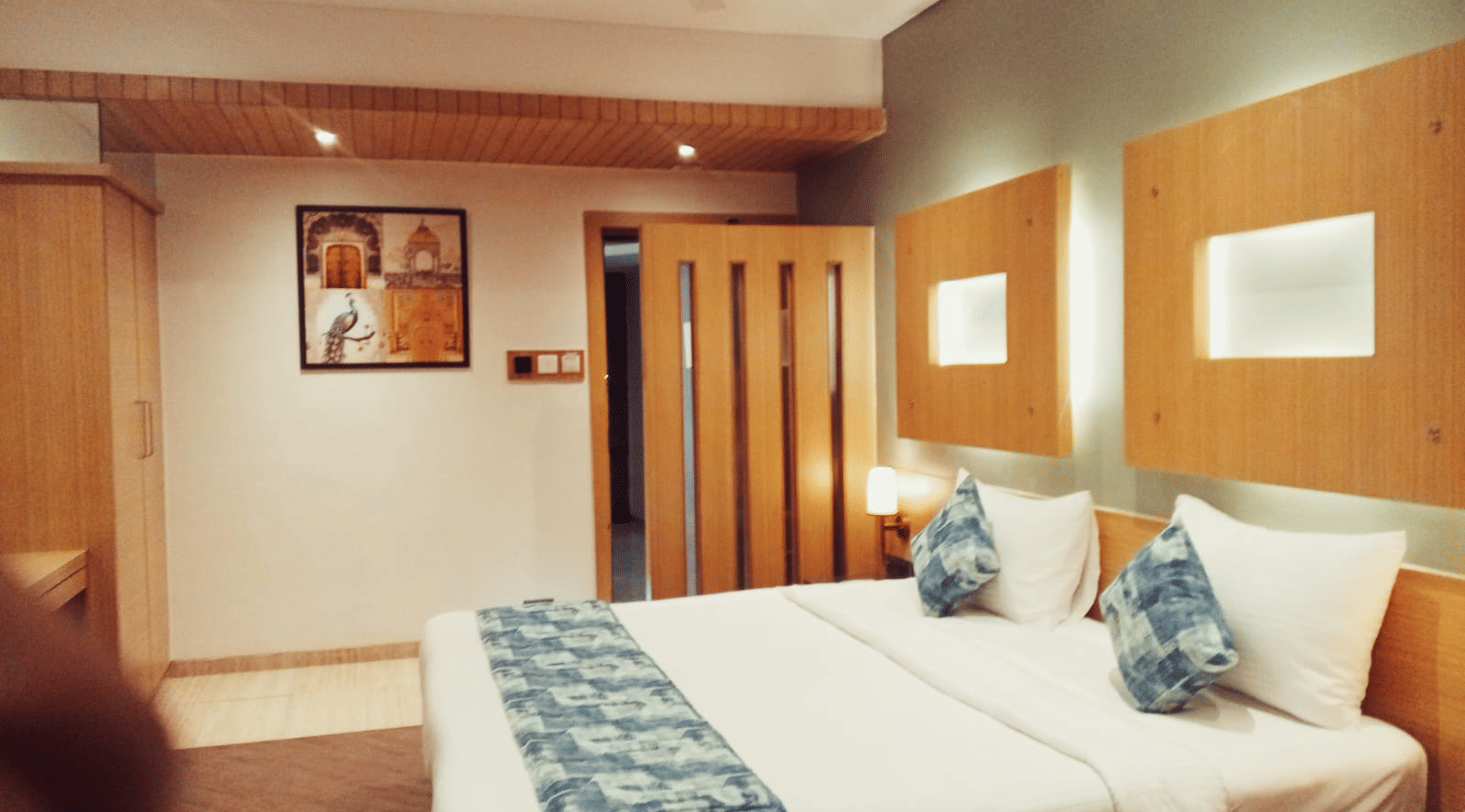 A bedroom space arranged with double beds, a wall-mounted wooden frame, an overhead ceiling fan, and a light-toned wooden wardrobe unit at Hotel Mumbai House, Thane.