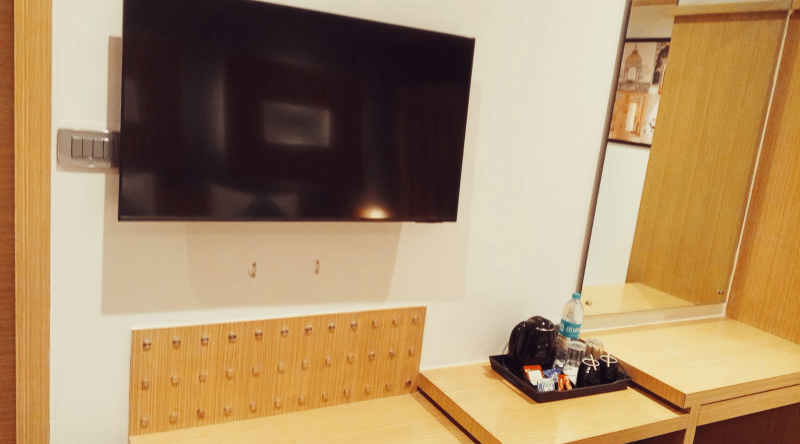 A flat screen television mounted on a white wall above a wooden luggage rack, a vanity mirror, and a matching console desk at Hotel Mumbai House, Thane.