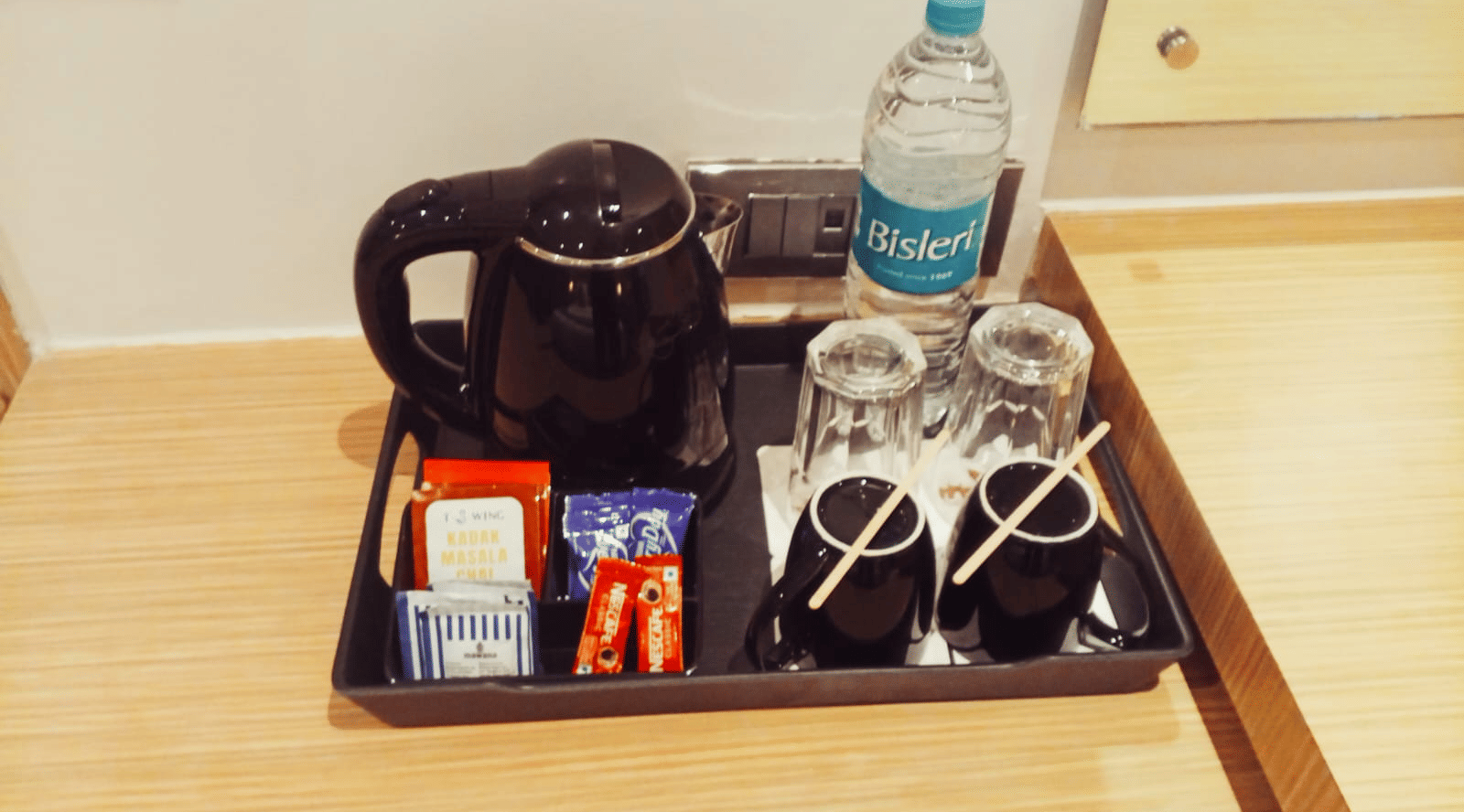 A black hospitality tray holding a black electric kettle, a clear plastic water bottle, 2 inverted glasses, and tea sachets on a wooden counter at Hotel Mumbai House, Thane.