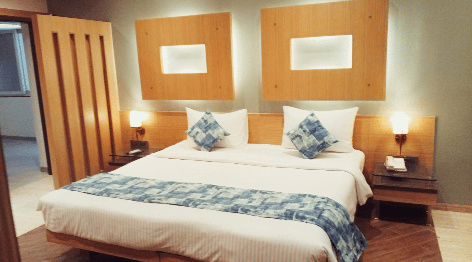 A king size bed with a white duvet, a patterned bed runner, 2 wooden nightstands with lamps, and 2 rectangular wall design panels at Hotel Mumbai House, Thane.
