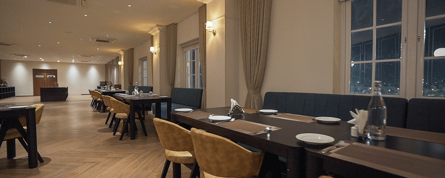 A hotels formal dining area at Pride Resort, Rajkot featuring long dark wooden tables, chairs, and comfortable booth seating nestled beneath large windows, ideal for group meals