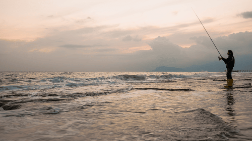 A person standing in shallow waters holding a fishing rod with ocean waves in the background.