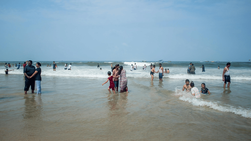 Crowds of people enjoying a paddle in the shallow waves of a broad, sandy beach under a bright, slightly hazy sky.