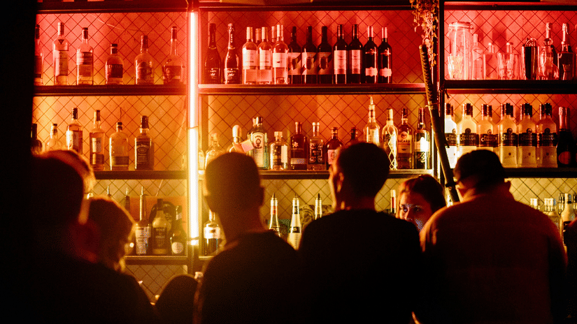 Guests gathered at a vibrant bar counter illuminated by warm lighting, with shelves of premium spirits creating a lively nightlife atmosphere
