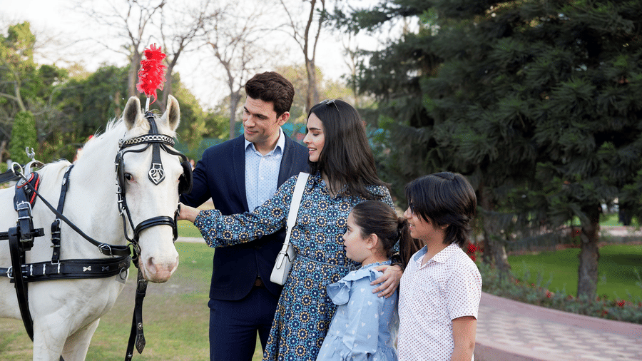 A family standing beside a horse with a red plume, petting it in a park with trees and a paved path nearby, at Heritage Village Resorts & Spa, Manesar.