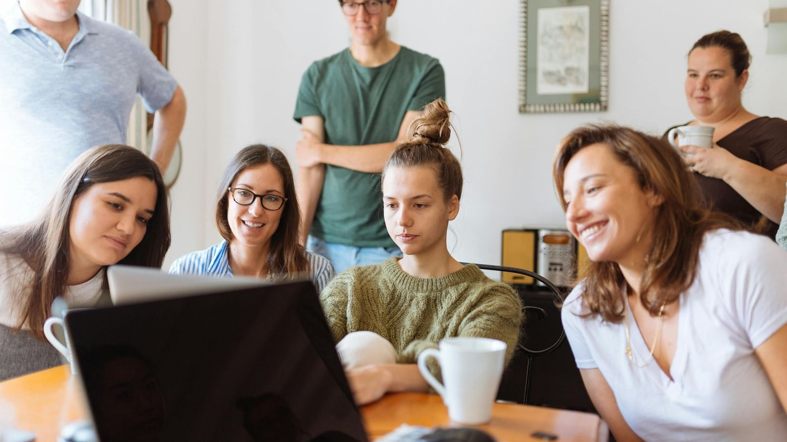A group of people are gathered around a laptop, with some looking at the screen and others standing behind them.