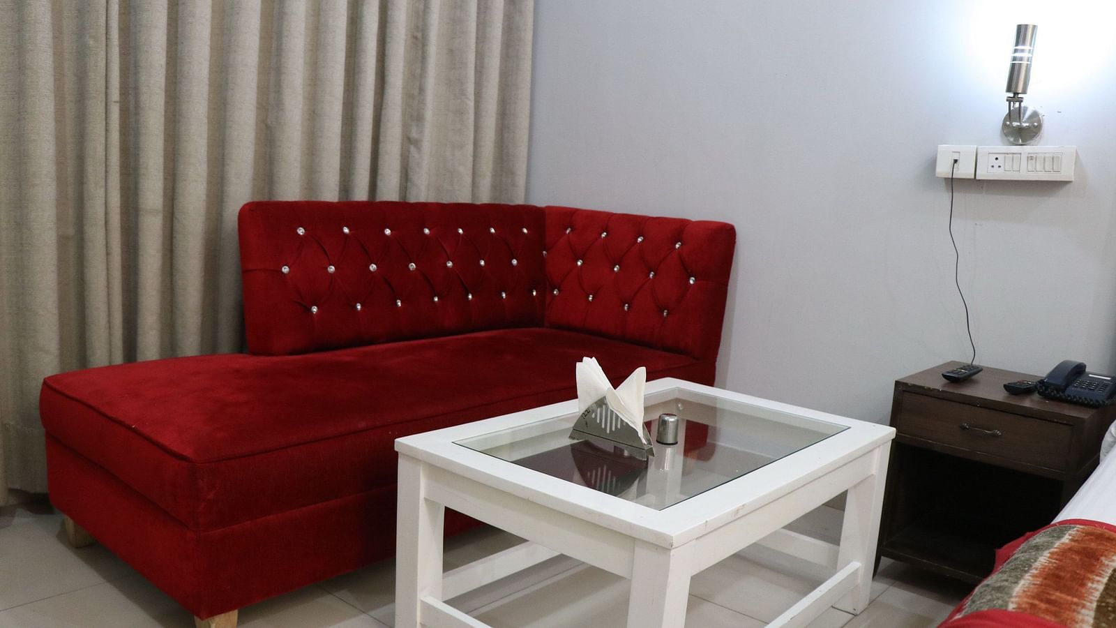A cosy corner with a red tufted sofa, a white square table, and a bedside table, illuminated by a wall lamp against a soft gray wall. - Hotel Diamond Plaza, Chandigarh.