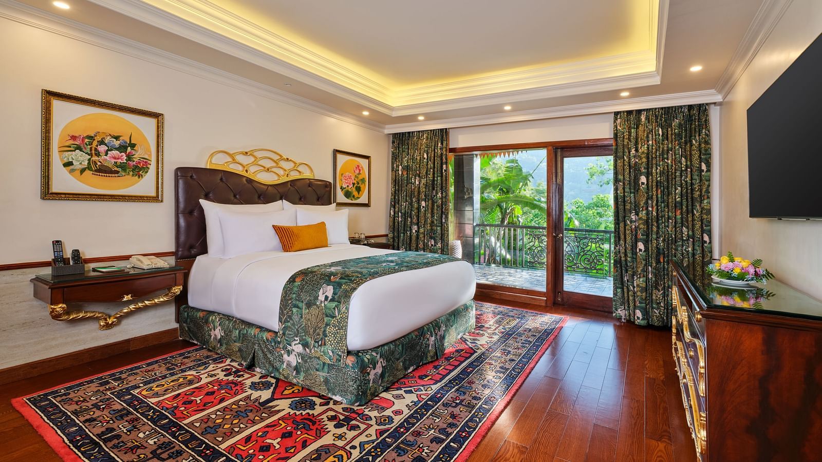 Luxurious Grand Villa bedroom with a large bed on a decorative rug, wooden cabinet, TV, artworks, and a window overlooking greenery .