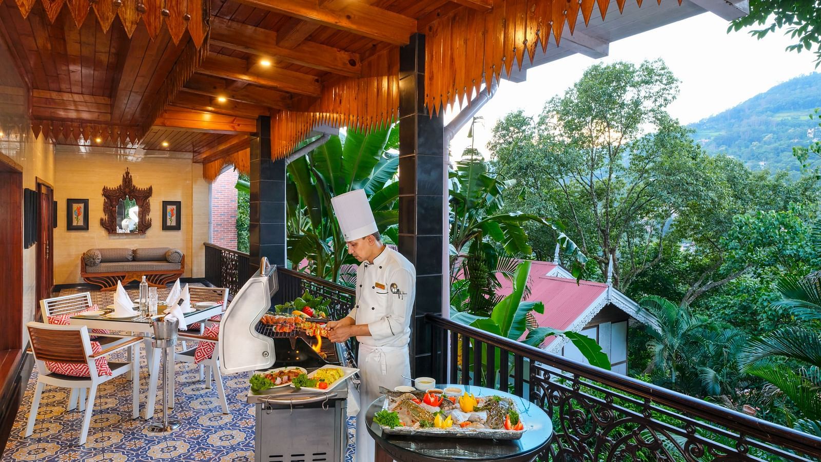 A chef in Grand Villa grilling food. The background has a seating area with tables, chairs, decorative tiles, wooden ceilings, and a view of outdoor greenery.