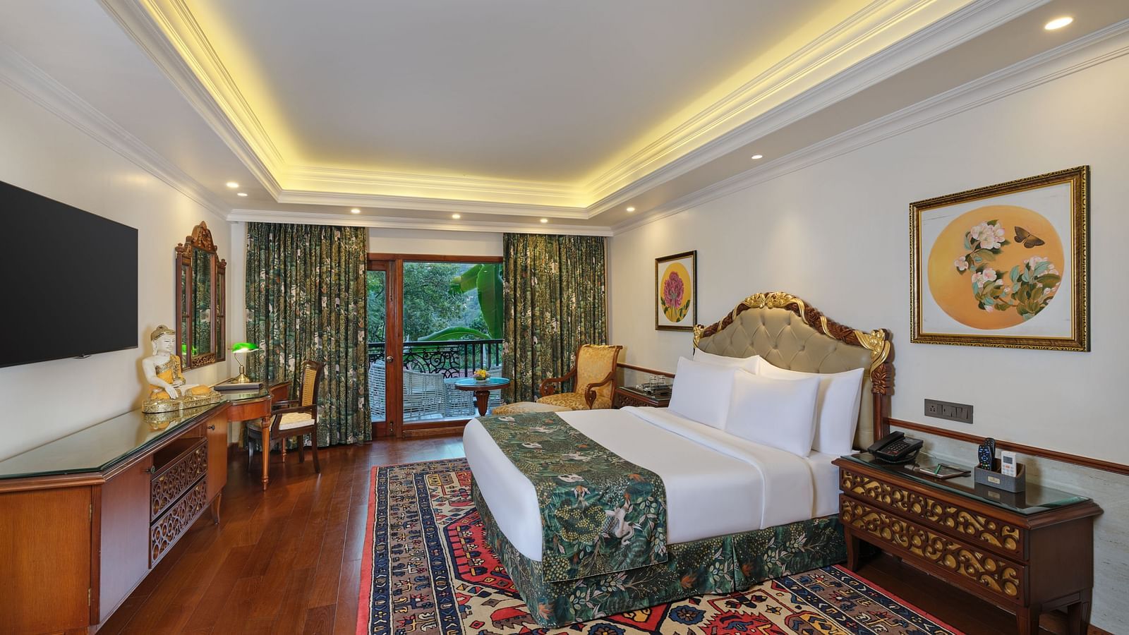 A luxurious Grand Villa bedroom with a floral comforter set, wooded nightstand, decorative rug, window overlooking greenery, TV, and framed artworks.