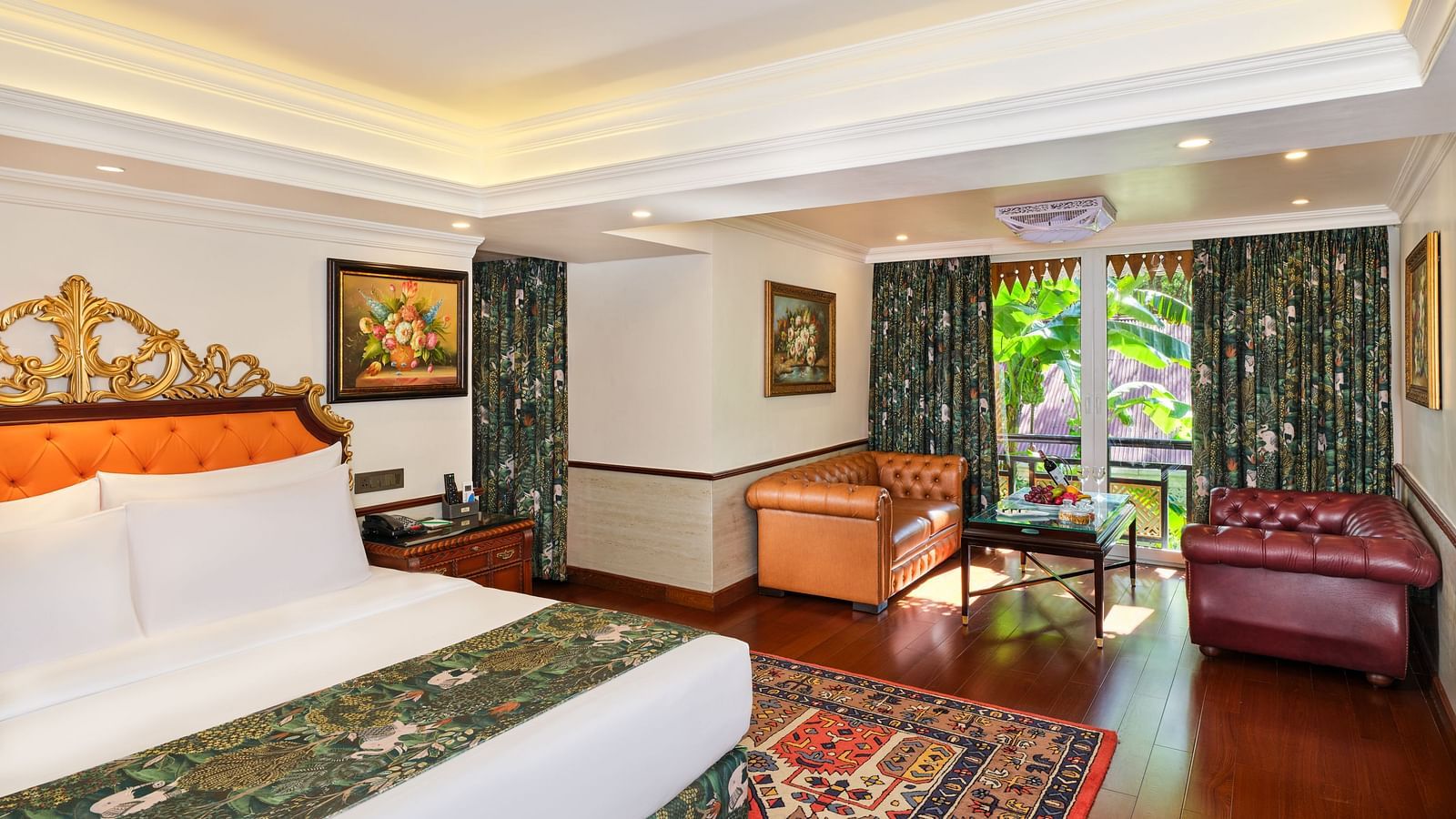 Oriental Villa bedroom with a grand bed on a decorative rug, artworks on wall, sofas and huge windows with curtains overlooking greenery.