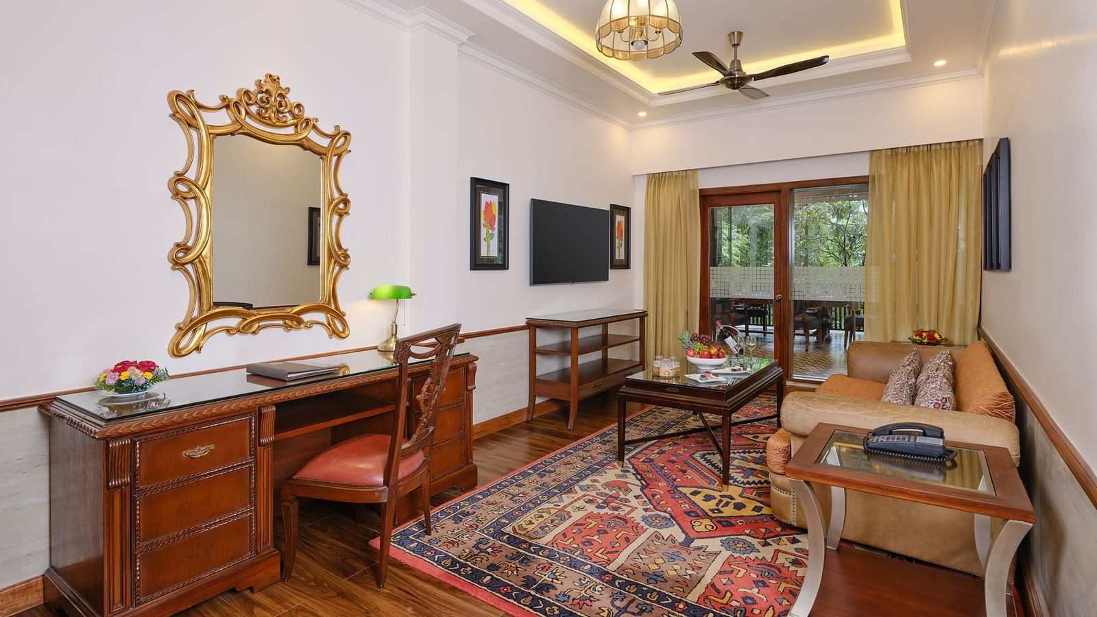 Interior seating area of Regent villa  with table, sofas, wooden cabinet, TV, mirror, and windows overlooking greenery.