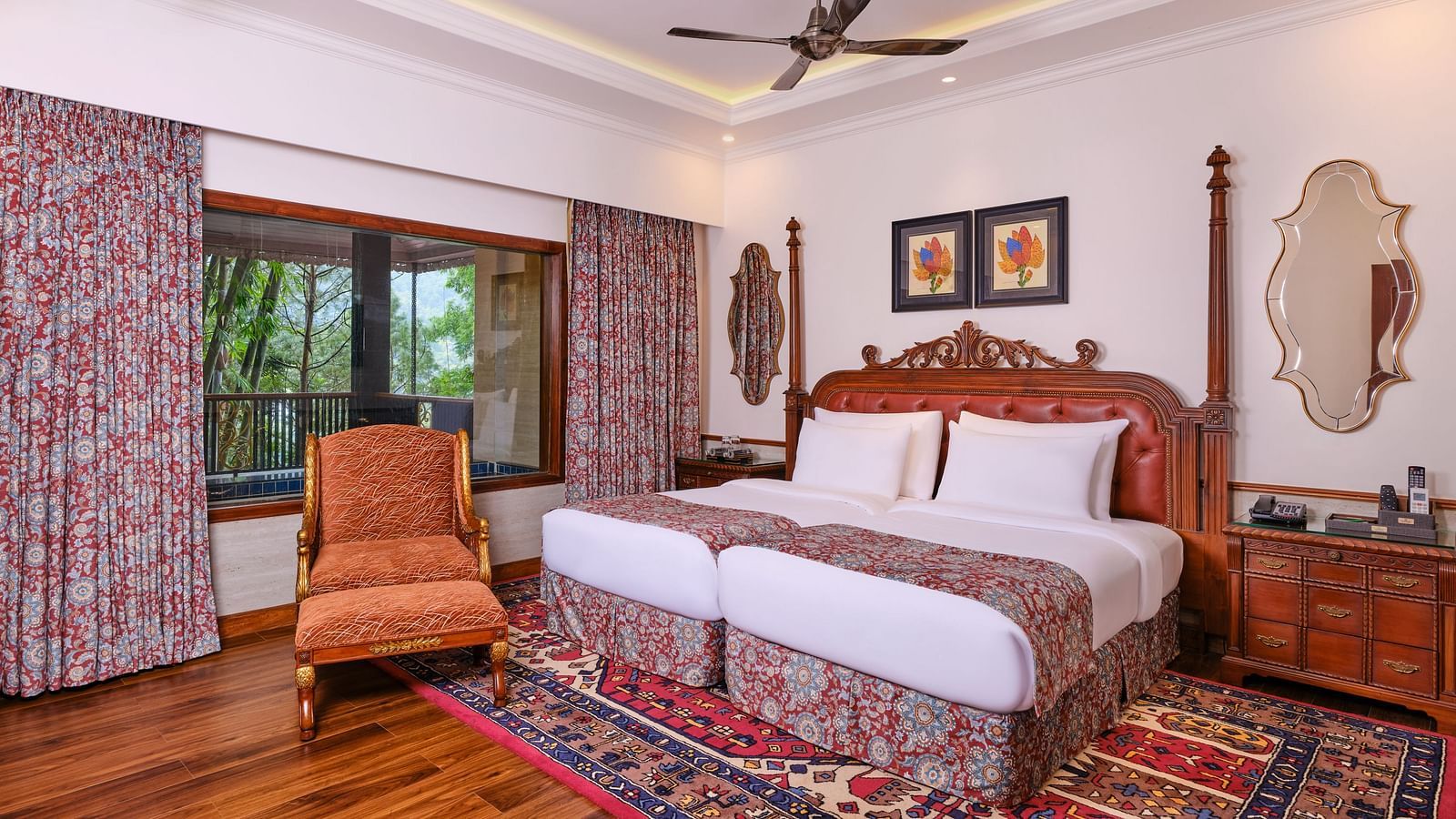 Regent villa bedroom with a huge bed on decorative rug, armchair, wooden cabinet, mirror and a window with curtains overlooking greenery.