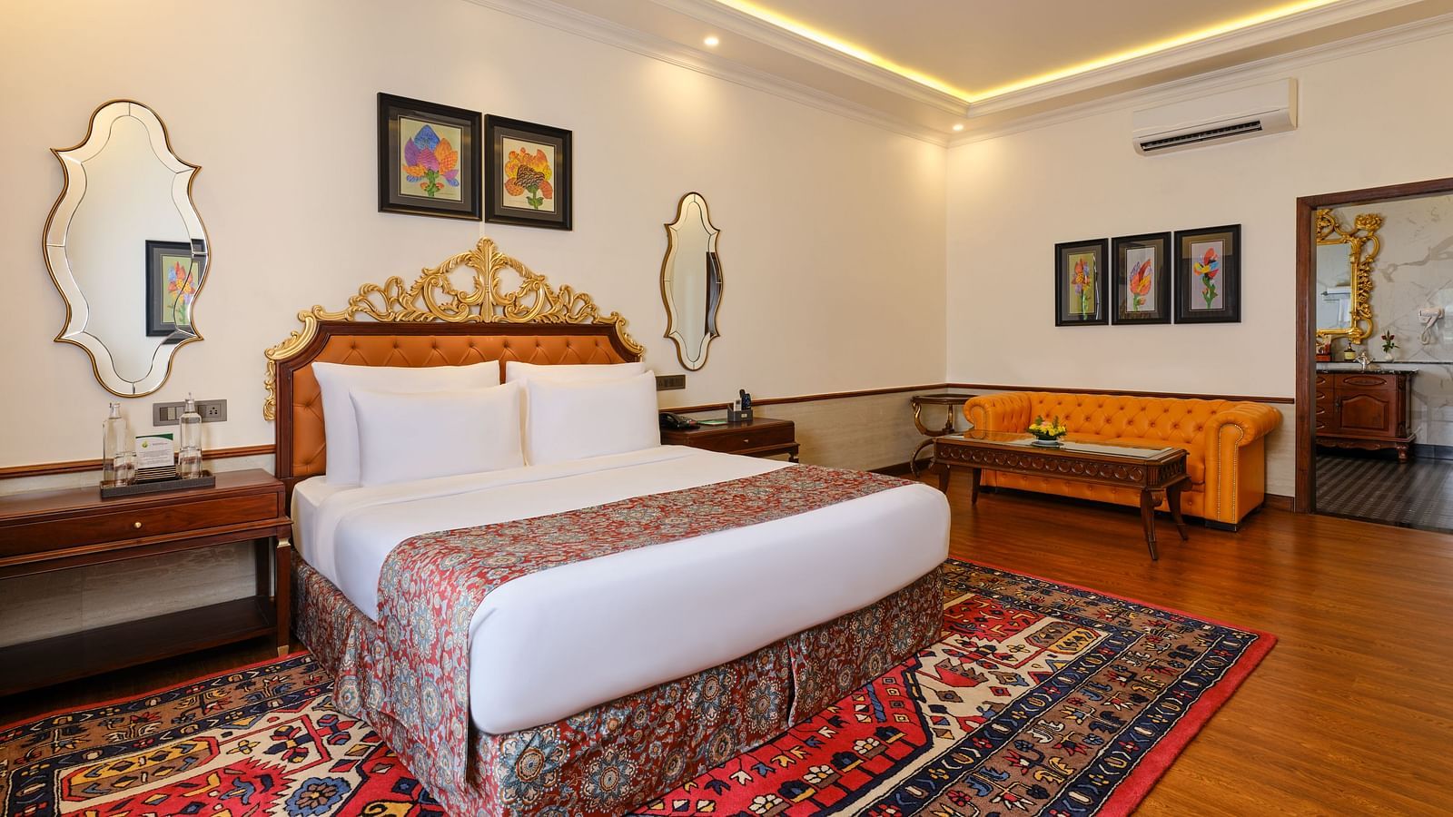 A luxurious suite, where a sofa, a table and a grand bed sits beneath a decorative headboard and artwork on the wall with two mirrors.