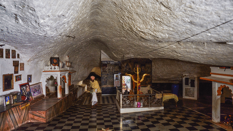 A checkered tile floor covers the ground inside a cave shrine containing altars, pictures, and a trident mounted on a platform surrounded by a fence.