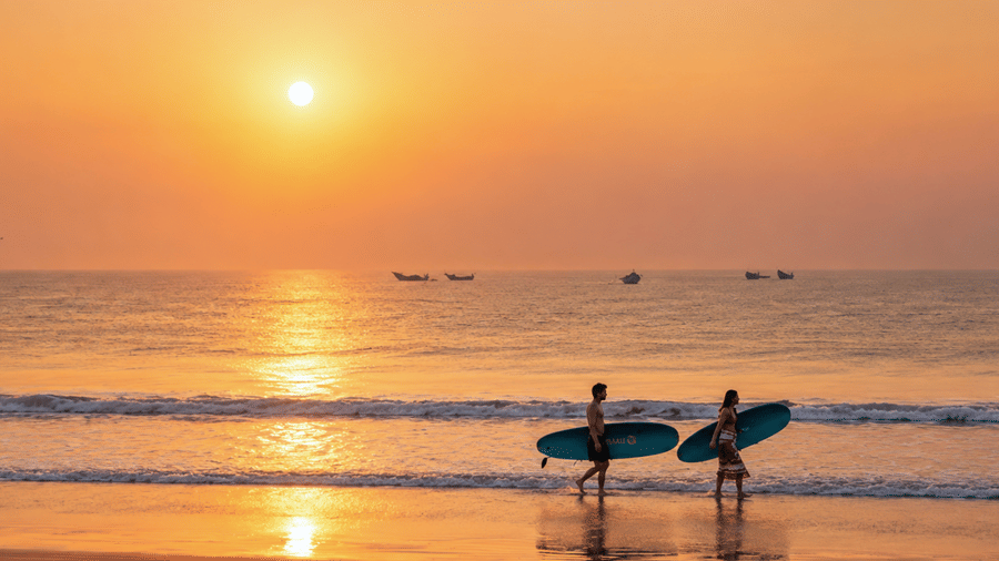 Two surfers carrying surfboards along the sandy beach during a bright sunrise at MAYFAIR Palm Beach Resort, Gopalpur-on-Sea. 