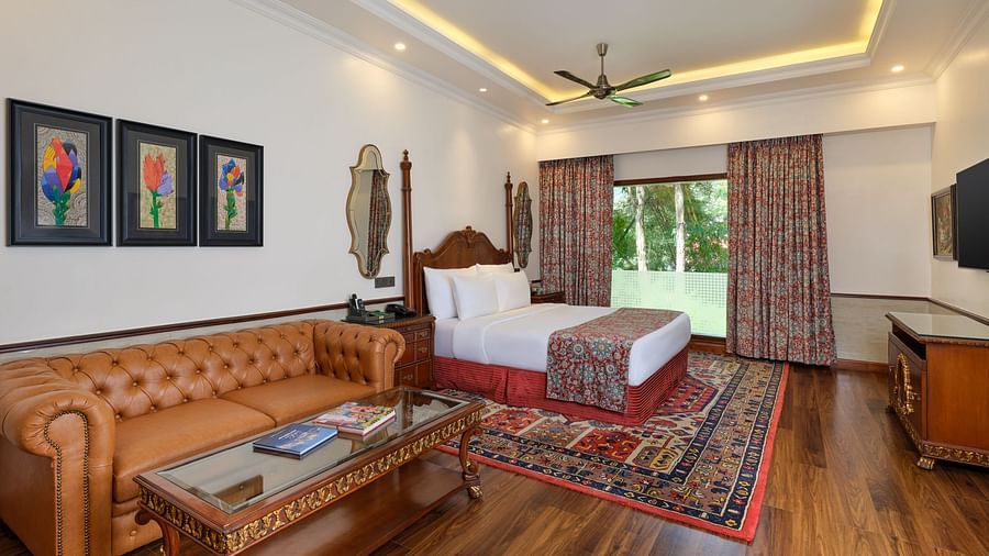 a Crown Villa bedroom with a large bed on a decorative rug, sofa, wooden cabinet, TV, artworks, and a window overlooking greenery  