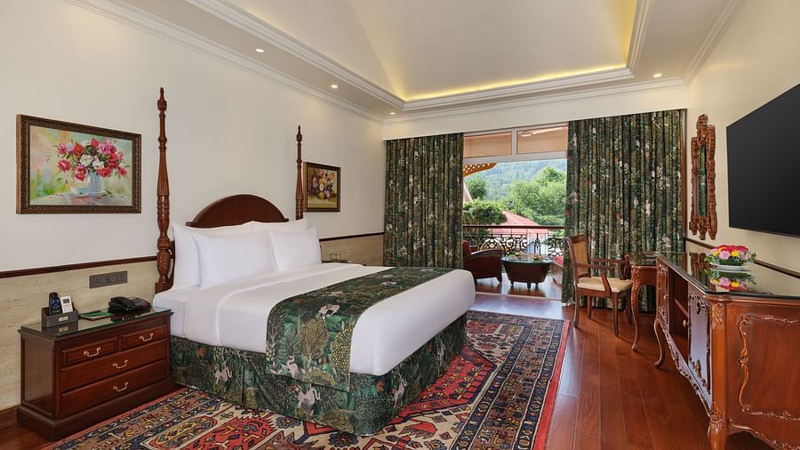 a Deluxe bedroom with a large bed on a decorative rug, wooden cabinet, TV, artworks, and a window overlooking greenery 
