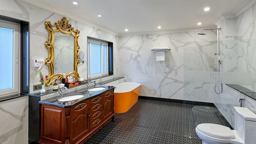 Luxurious Executive Suite Bathroom with a large mirror above a wooden cupboard with two sinks. An orange Bathtub, a commode  and a walk in shower are set against marble walls