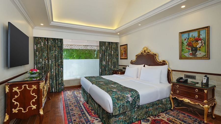 a luxurious Family Room bedroom with a large bed and floral comforter set, wooded nightstand, decorative rug, window overlooking greenery, TV, and framed artworks