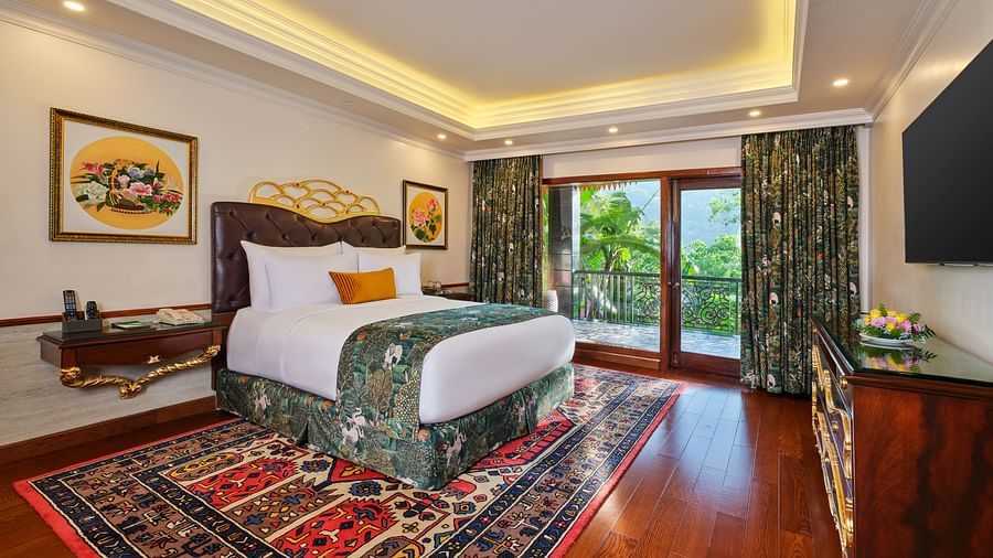 luxurious Grand Villa bedroom with a large bed on a decorative rug, wooden cabinet, TV, artworks, and a window overlooking greenery 
