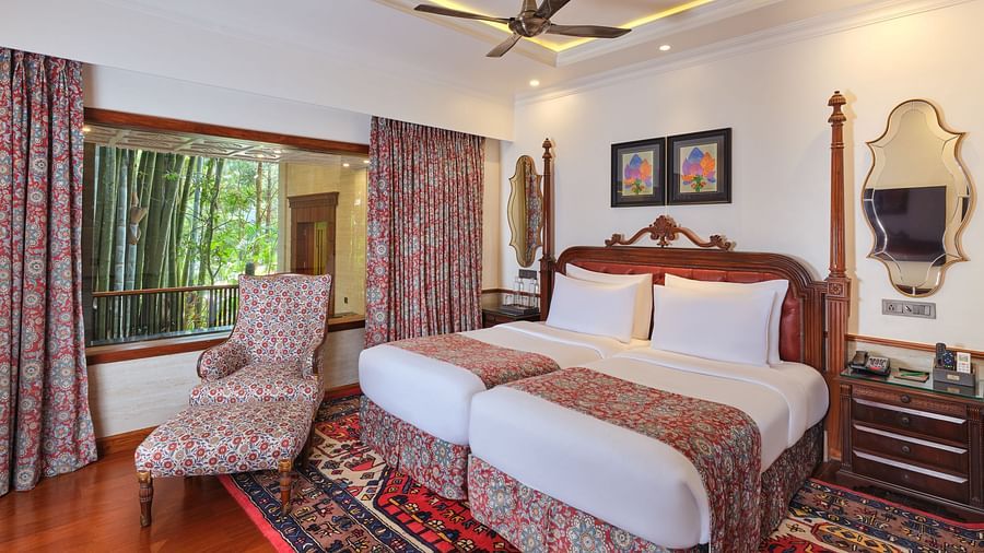 Royal Villa featuring a wooden bed with white bedding, a patterned armchair, a decorative rug, a ceiling fan, framed pictures, and a window with bamboo trees, complemented by a patterned curtain