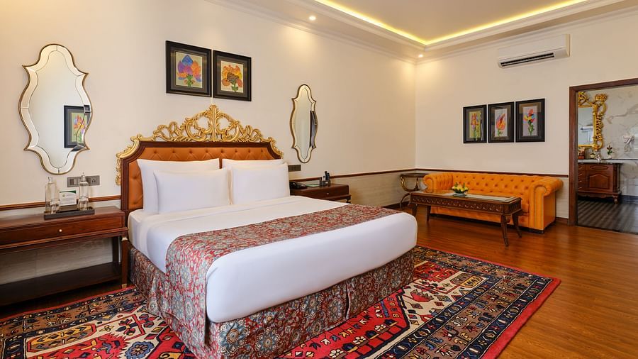 a luxurious suite, where a sofa, a table and a grand bed sits beneath a decorative headboard and artwork on the wall with two mirrors