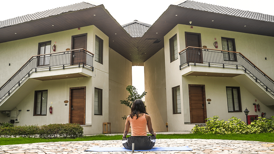 A person practicing yoga on a mat in a serene outdoor setting, with resort buildings in the background - Shervani Pebbles & Pines, Corbett