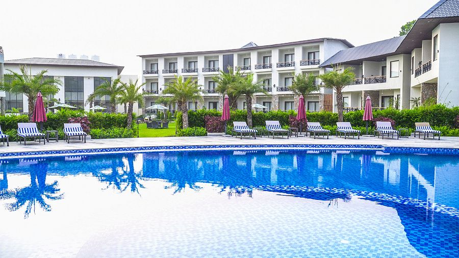 A refreshing outdoor pool surrounded by sunbeds with umbrellas, offering a  view of the resort's lush garden and modern architecture - Shervani Pebbles & Pines, Corbett
