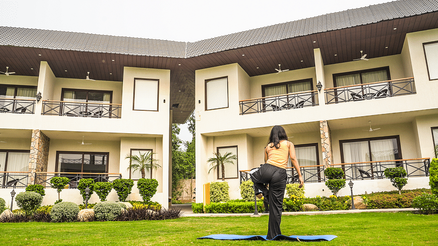 A woman practising yoga on a mat in a lush garden, with a scenic view of a resort building in the background - Shervani Pebbles & Pines, Corbett