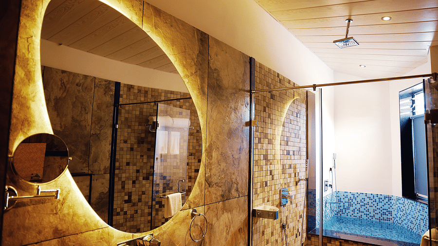 A bathroom of Luxury Villa with Sitout featuring bathroom mirror with a unique shape and backlighting, along with a glimpse of a bed and blue armchair in the background at The Qualia Resort Club & Brewery, Udaipur.