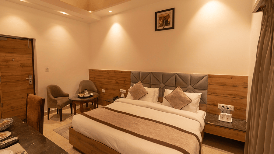 A warm and inviting resort suite featuring a double bed with upholstered headboard, wooden accents, and a small sitting area - Wood Castle Spa & Resort, Jim Corbett