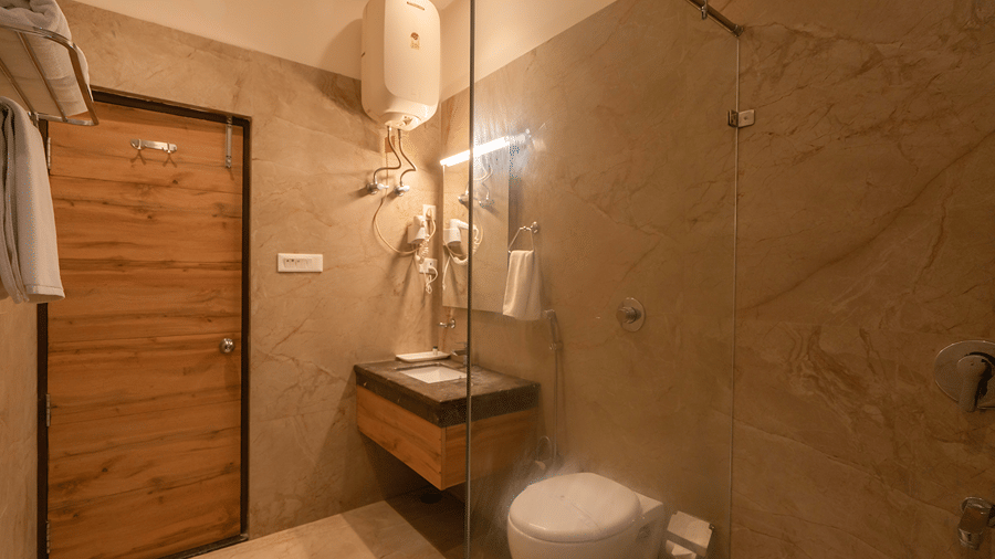 A modern en-suite bathroom with beige tiles, a glass shower partition, wall-hung toilet, and wooden door - Wood Castle Spa & Resort, Jim Corbett