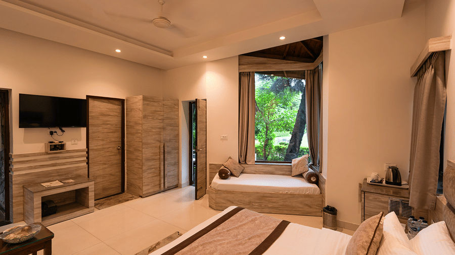 A spacious luxury resort bedroom featuring a plush double bed, wall-mounted TV, and a cosy window daybed seating area - Wood Castle Spa & Resort, Jim Corbett
