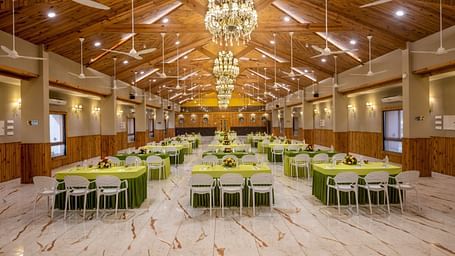 A well-lit banquet hall with multiple tables and chairs set for an event at Angel Lords Eco Inn Amreli.