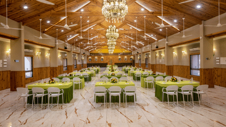 A well-lit banquet hall with multiple tables and chairs set for an event at Angel Lords Eco Inn Amreli.