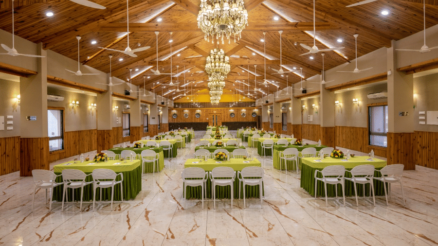 A well-lit banquet hall with multiple tables and chairs set for an event at Angel Lords Eco Inn Amreli.