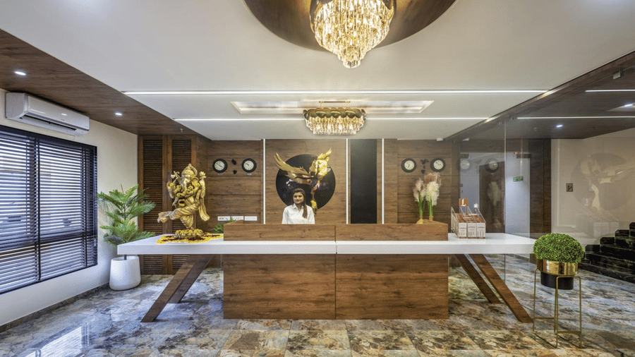 A well-lit hotel reception area with a wooden front desk and chandelier at Angel Lords Eco Inn Amreli.