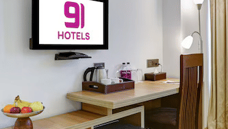 A close-up of the room's amenities including a wooden desk, a wall-mounted TV with branding, a mini-fridge, and a tray of refreshments - Hotel 91 HUDA City Centre, Gurugram