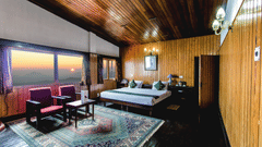 An image of a cosy room with warm wooden interiors, featuring a neatly arranged bed, sofa chairs, and a table, complemented by large glass windows offering a beautiful view of the mountains and the sun - Central Gleneagles Heritage Resort, The Mall Road, D