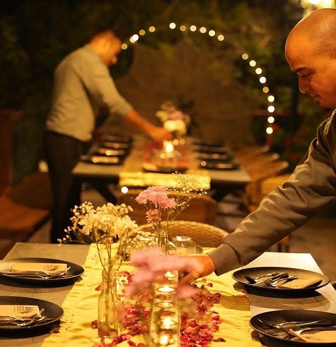 Dining table at Karma Chalets set with plates and a flower garland, with a person arranging the setup under lights.