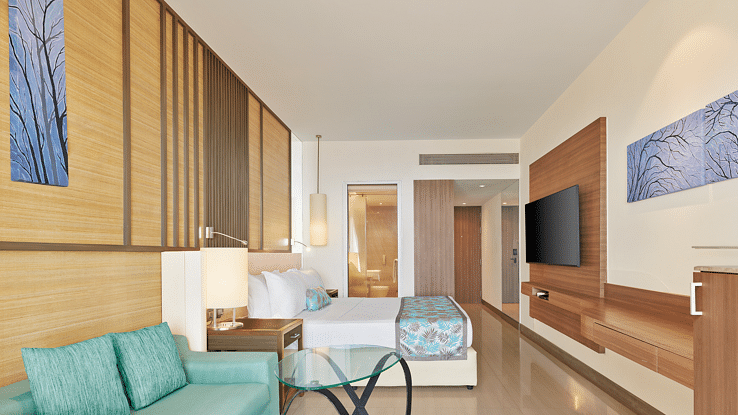 Stylish modern room with wooden accents, pastel furnishings, a wall-mounted TV and an open layout leading to a spacious ensuite – The Resort, Mumbai.
