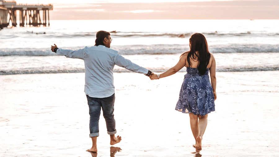 A couple holding hands and walking on the shoreline, basking in the beauty of a sunset over the ocean.
