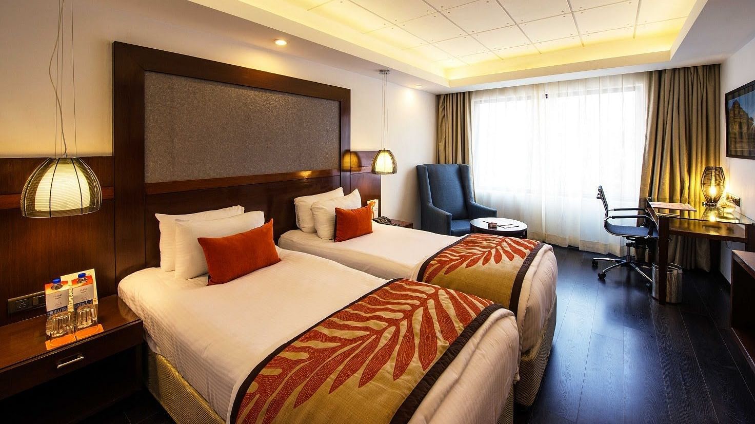 Deluxe Room at The Cunningham Hotel featuring twin beds with decorative runners, warm lighting, pendant lamps, a seating area, and a functional workspace near a large window with natural light