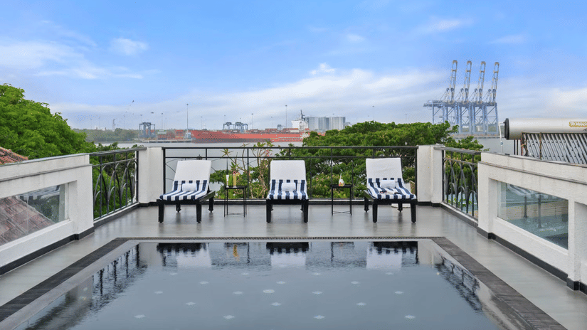Rooftop swimming pool deck at The Old Court House by Abad, one of the luxury hotels with swimming pool in Kochi, showcasing three striped lounge chairs, glass railings, and a scenic view of the harbour under a bright sky.
