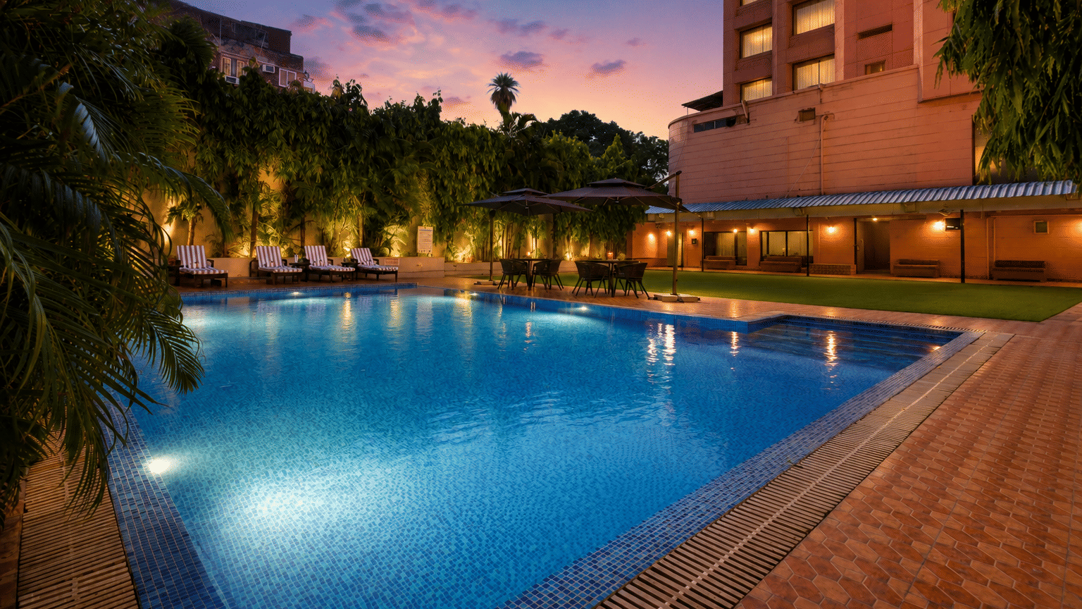Illuminated hotel pool at dusk with lounge chairs, tropical greenery, and warm ambient lighting.